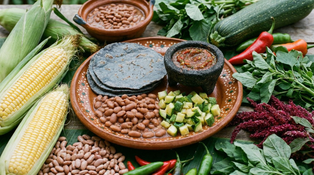 Un plato tradicional mexicano con tortillas de maíz azul, frijoles de la olla, calabacitas picadas y una salsa de chile molcajeteada, rodeado de ingredientes frescos de la milpa
