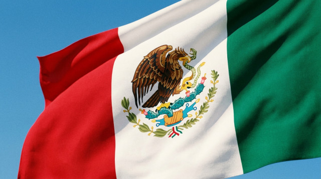 Primer plano de la Bandera de México ondeando majestuosamente con el Escudo Nacional detallado en el centro bajo un cielo azul despejado