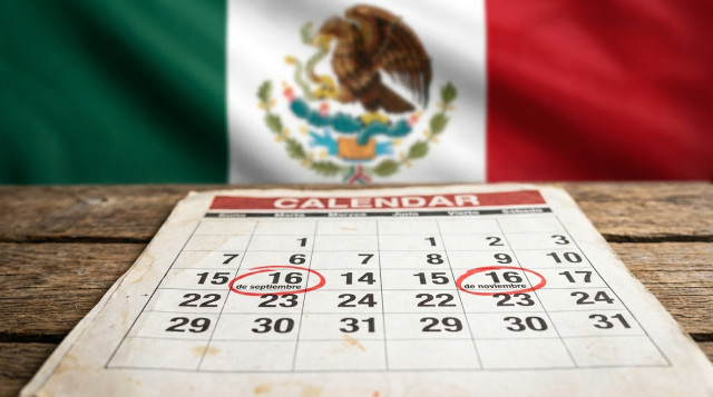 calendario impreso sobre una mesa con marcadores rojos en 16 de septiembre y 16 de noviembre, fondo con bandera mexicana desenfocada, foco en las fechas; texto alternativo 'Curiosi