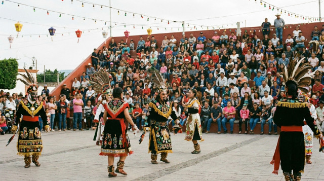 Fotografía horizontal de una plaza municipal durante una fiesta local en México: danzantes con trajes tradicionales en primer plano, público variado en graderío al fondo, faroles y