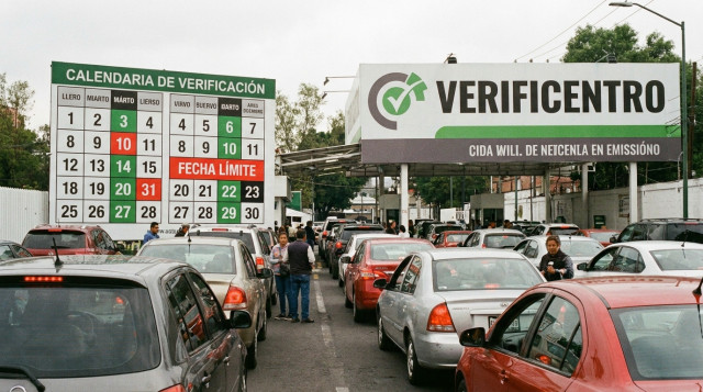 Línea de automóviles esperando inspección de emisiones en un verificentro oficial con el calendario de verificación vehicular visible en la señalética